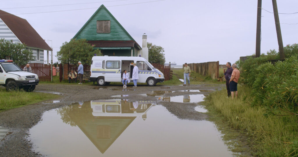 Zdjęcie przedstawiające karetkę pogotowia, przy niej stoją lekarze, wokół także inni ludzie. Za samochodem znajdują się domy.