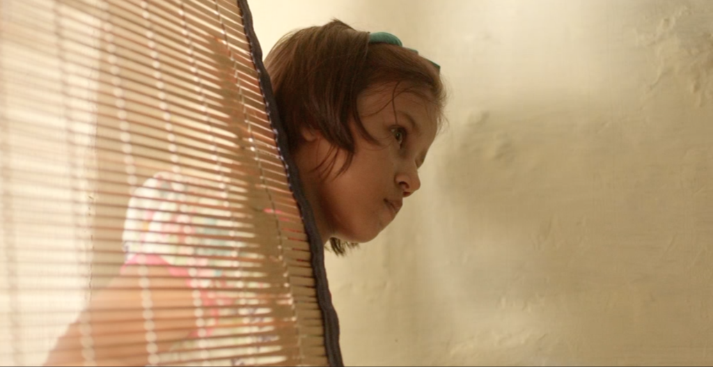 Zdjęcie przedstawiające małą dziewczynkę wyglądającą zza słomianej rolety.