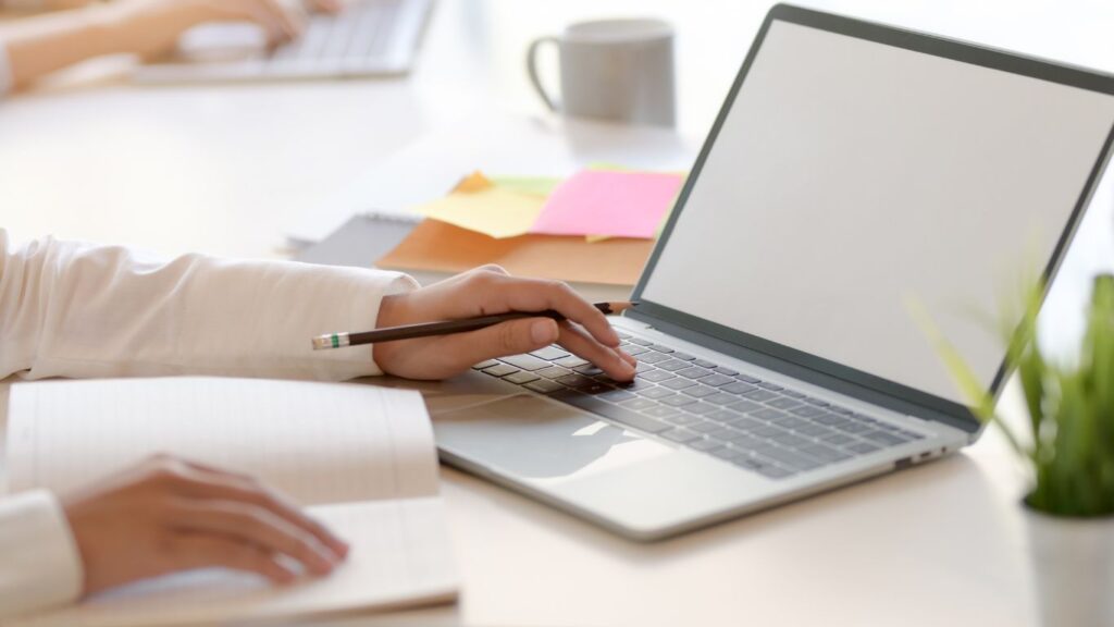 Zdjęcie biurka, na nim laptop i pracująca na nim dłoń trzymająca ołówek.