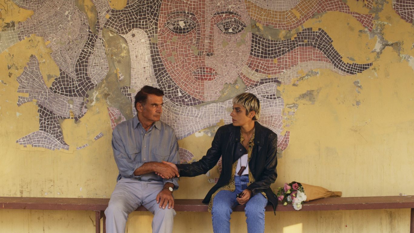 MOKRY PIASEK (2021), reż. Elene Naveriani, fabularny, 115', GRUZJA, SZWAJCARIA. Zdjęcie przedstawia młodą kobietę i starszego mężczyznę podających sobie dłonie. Siedzą na tle mozaiki.