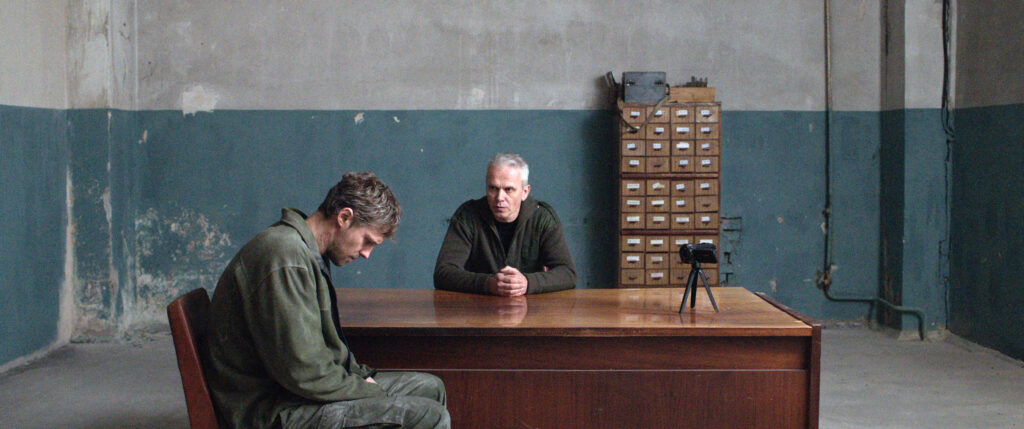 Zdjęcie przedstawiające dwóch mężczyzn siedzących przy biurku. Jeden z nich jest przesłuchiwany.