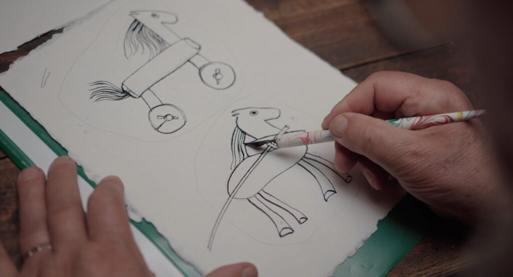 Zdjęcie przedstawiające kartkę oraz dłoń, która rysuje na niej piórem konia.