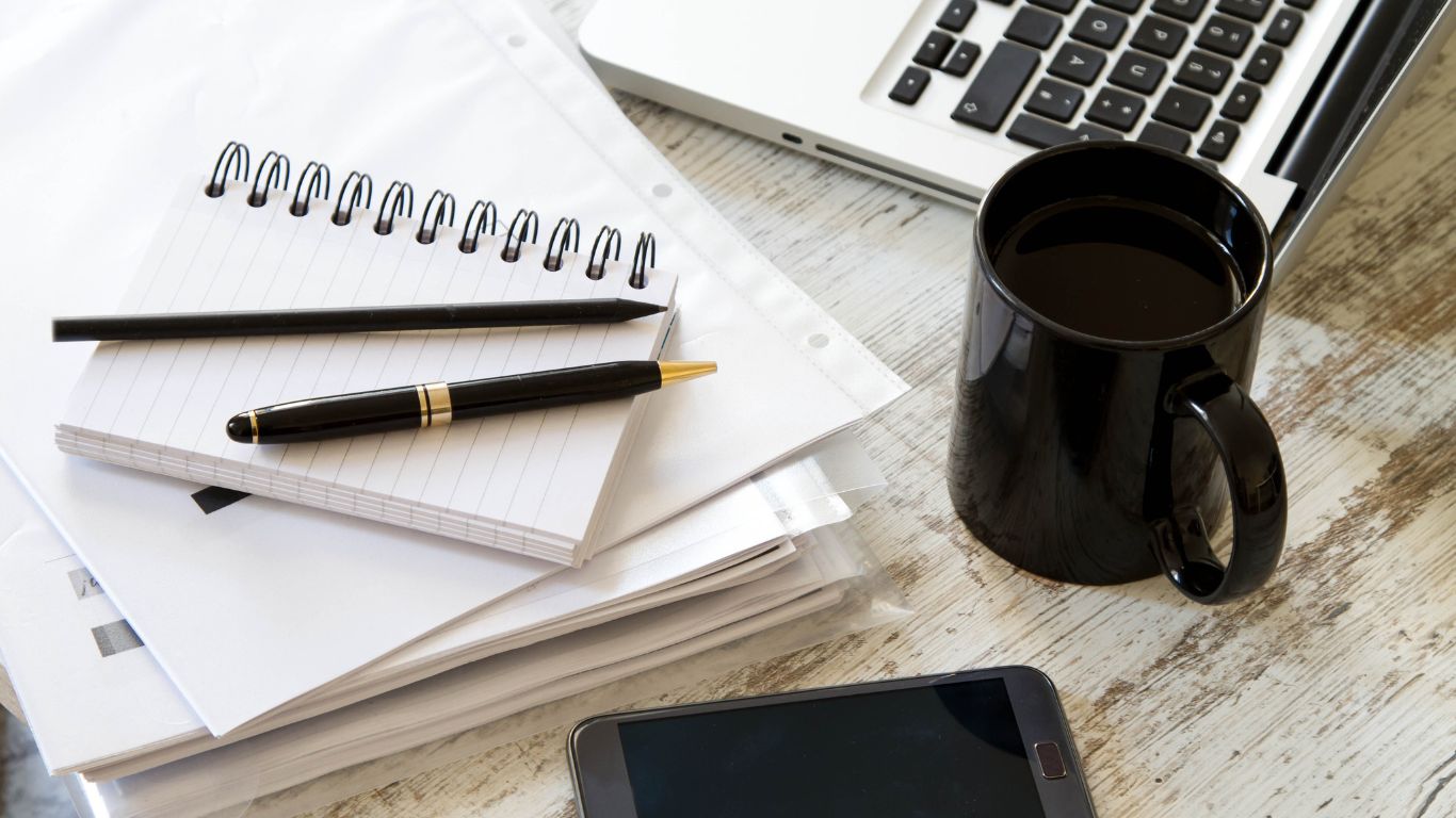 Zdjęcie biurka, na nim notes, ołówek, długopis, klawiatura laptopa, telefon i kubek z kawą.