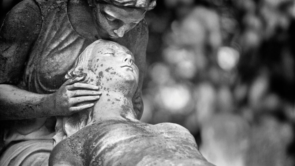 Biało czarne zdjęcie rzeźb cmentarnych. Kobieta całuje w czoło leżącą na jej kolanach zmarłą kobietę